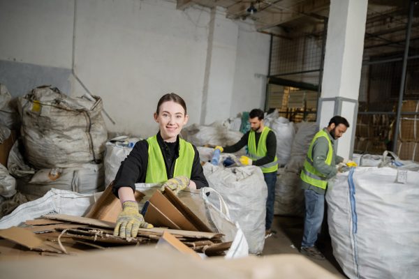 worker in gloves and high visibility vest looking at camera while putting cardboard in sack and working near multiethnic colleagues in waste disposal station, garbage sorting and recycling concept